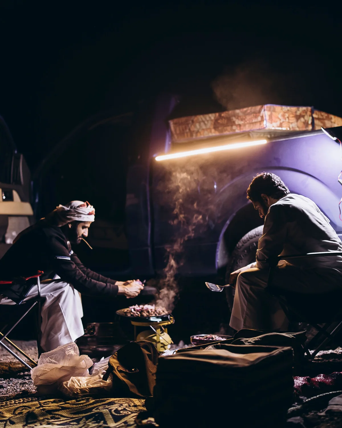 Two men cook on a portable gas stove beside their car at night, with smoke rising from the pan