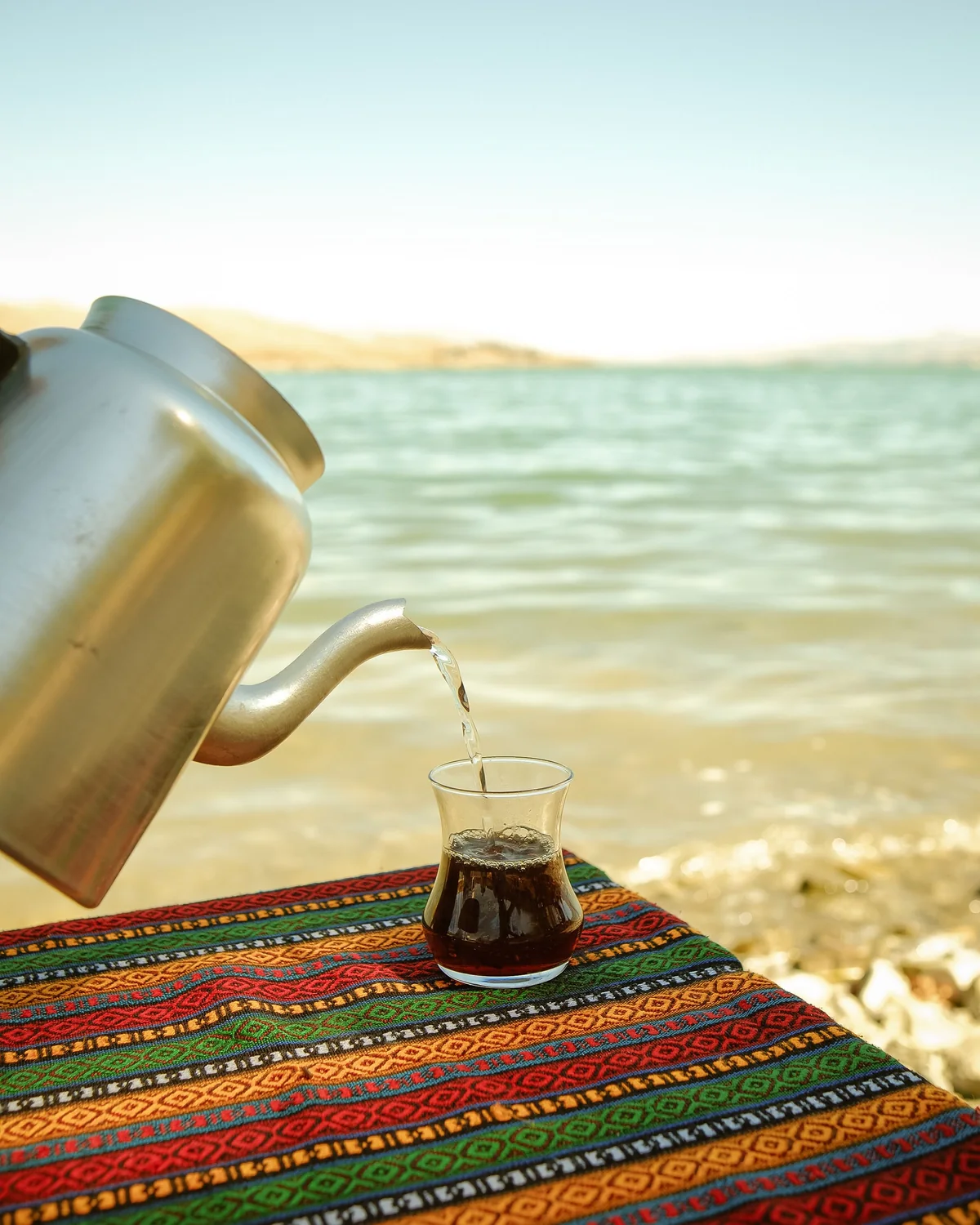 Tea poured from a metal kettle into a small glass, on a woven cloth by the water
